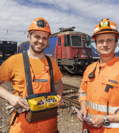 Zwei SBB-Carco-International-Mitarbeitende in stolzer Haltung und oranger Arbeitskleidung vor einer SBB-Cargo-Lok
